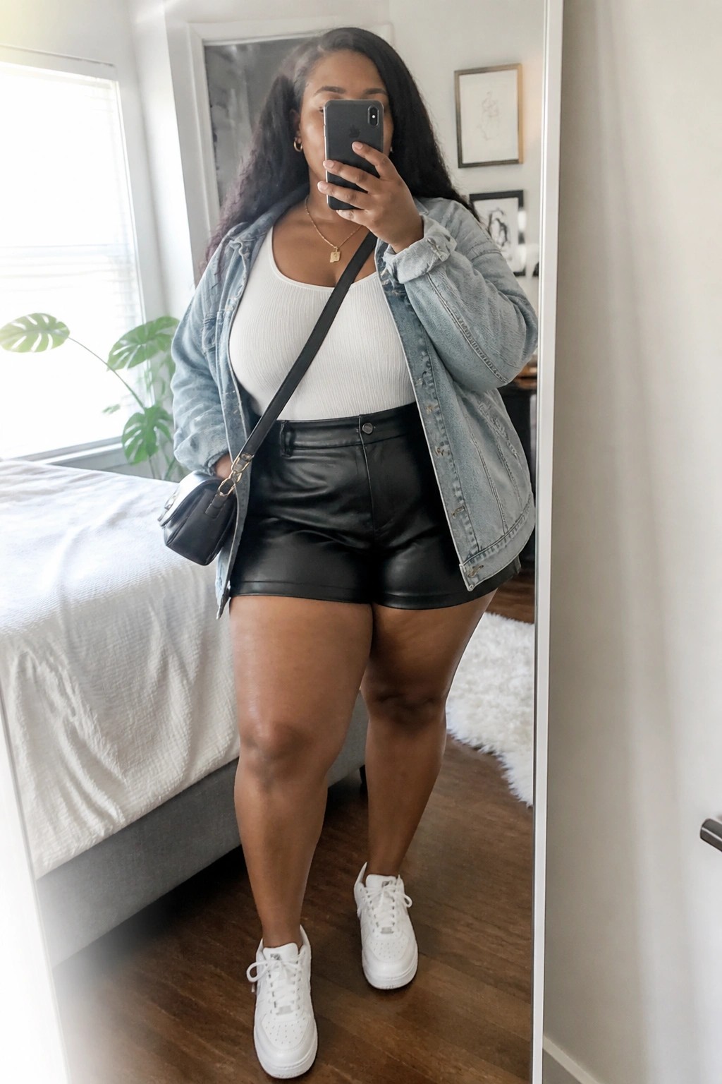 Woman in denim jacket and black leather shorts taking bedroom mirror selfie