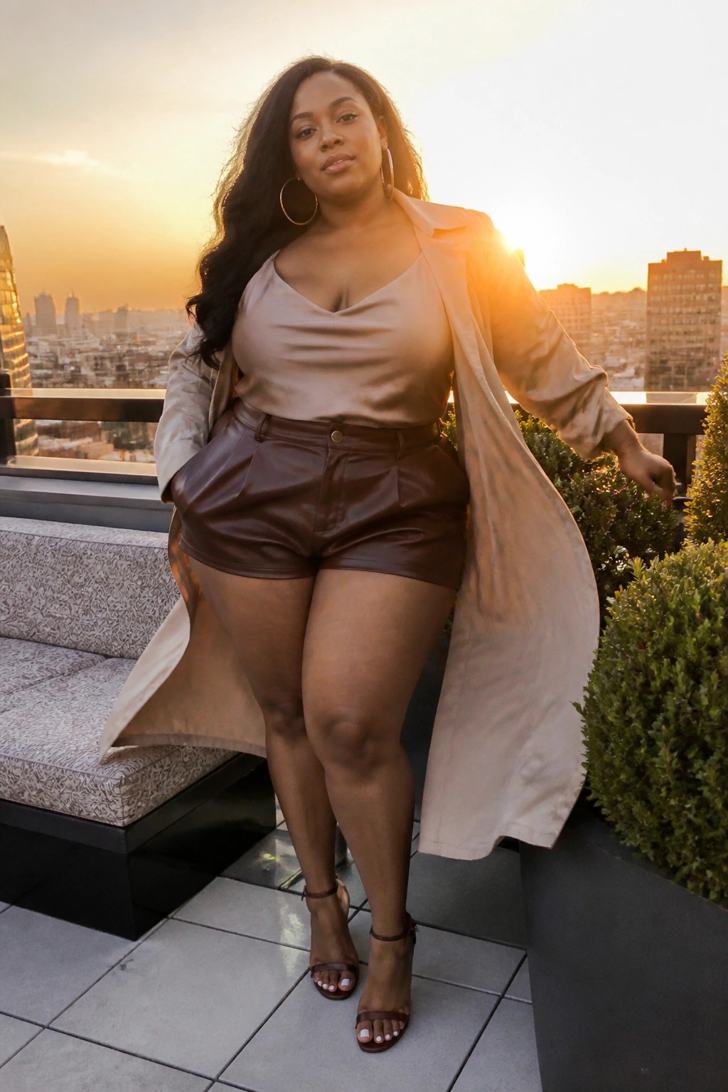 Woman in beige trench coat and brown leather shorts on sunset rooftop balcony