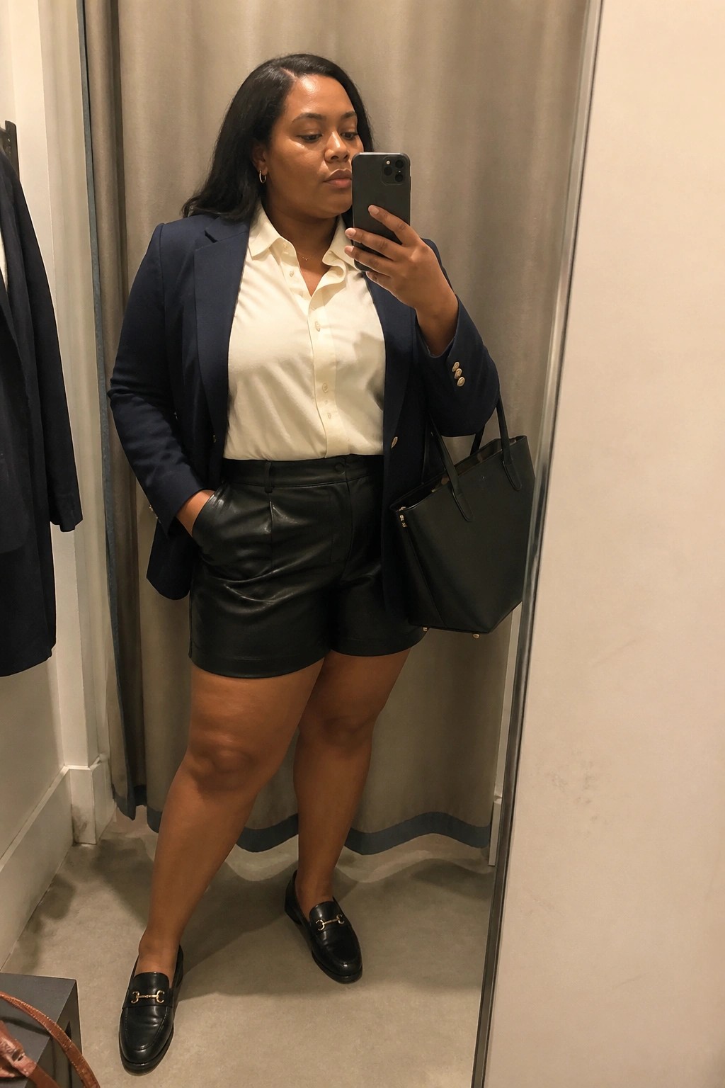 Woman in navy blazer and black leather shorts taking mirror selfie in fitting room