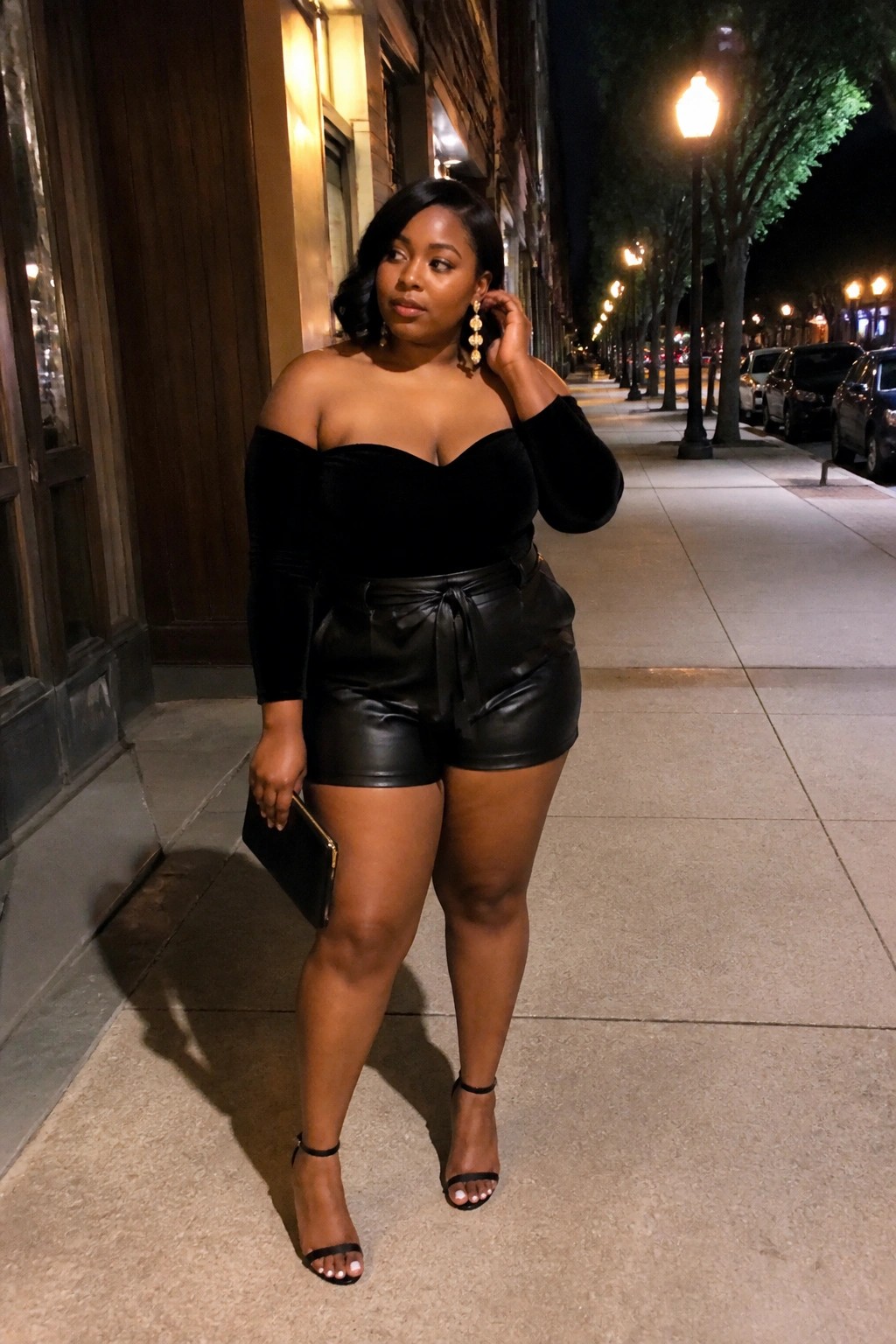 Plus size Black woman in black off shoulder velvet top with one long sleeve black leather shorts with tie belt black strappy heels and gold earrings holding a black clutch on a sidewalk.