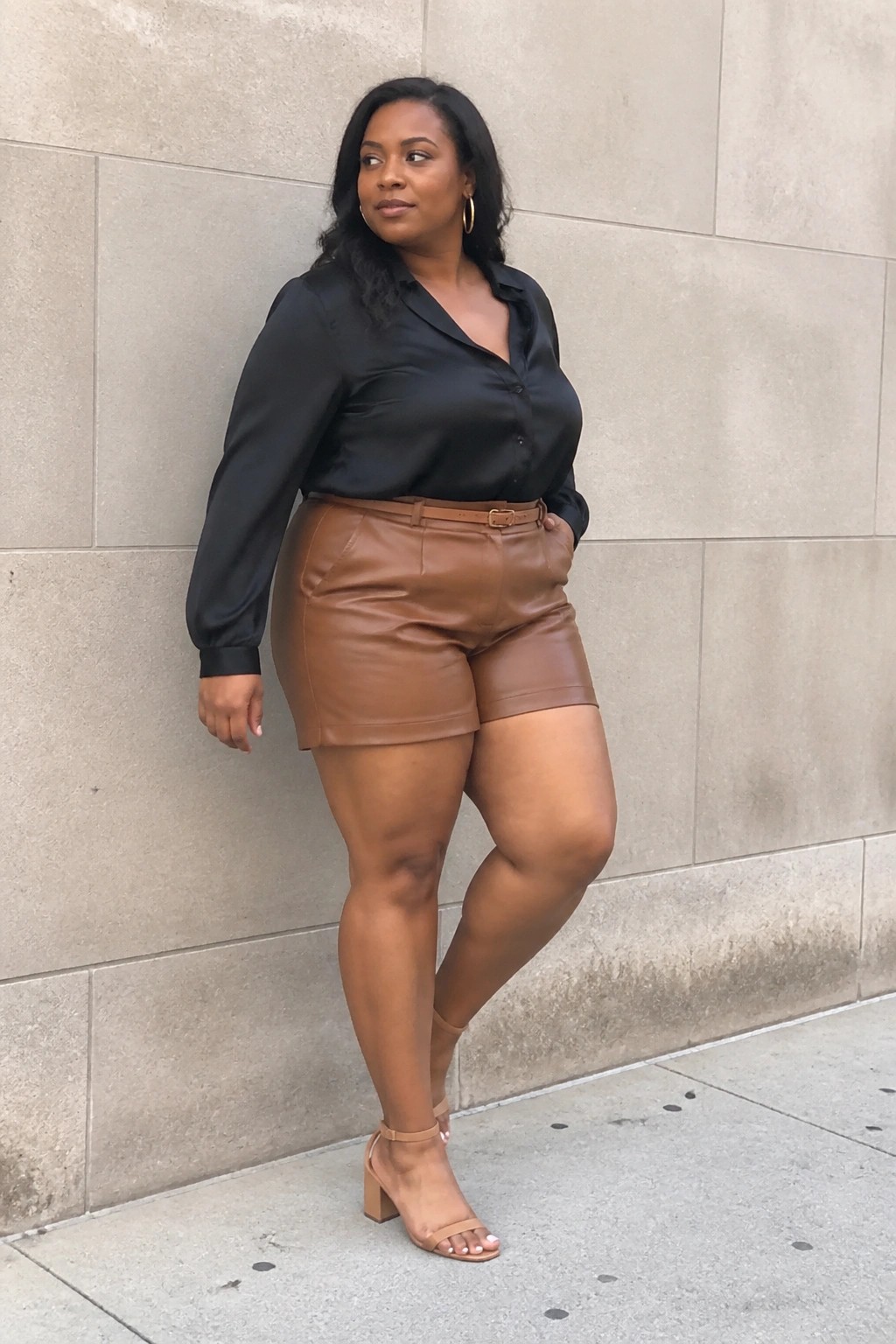 Woman in black satin blouse and brown leather shorts posing against stone wall