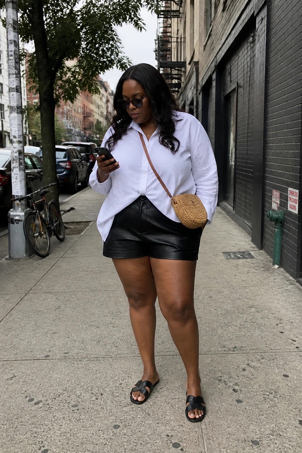 Plus size Black woman in an oversized white button up shirt and black leather shorts with a woven crossbody bag black slide sandals and sunglasses standing on a sidewalk.