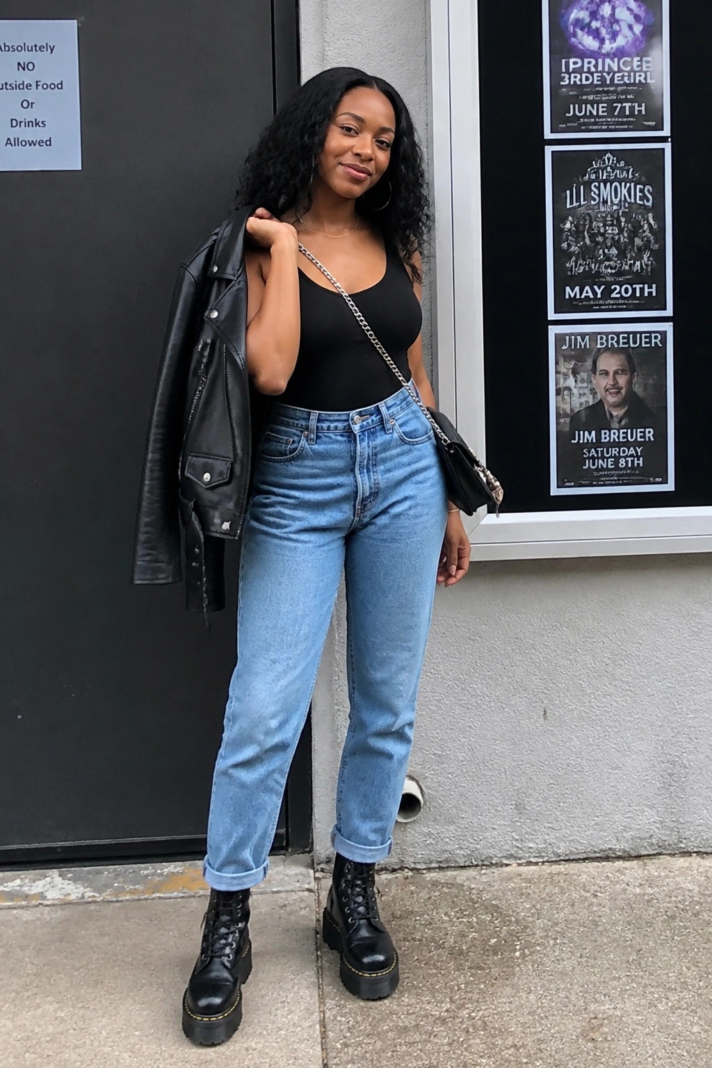 Curly-haired woman in black top and blue jeans holds leather jacket outside venue.