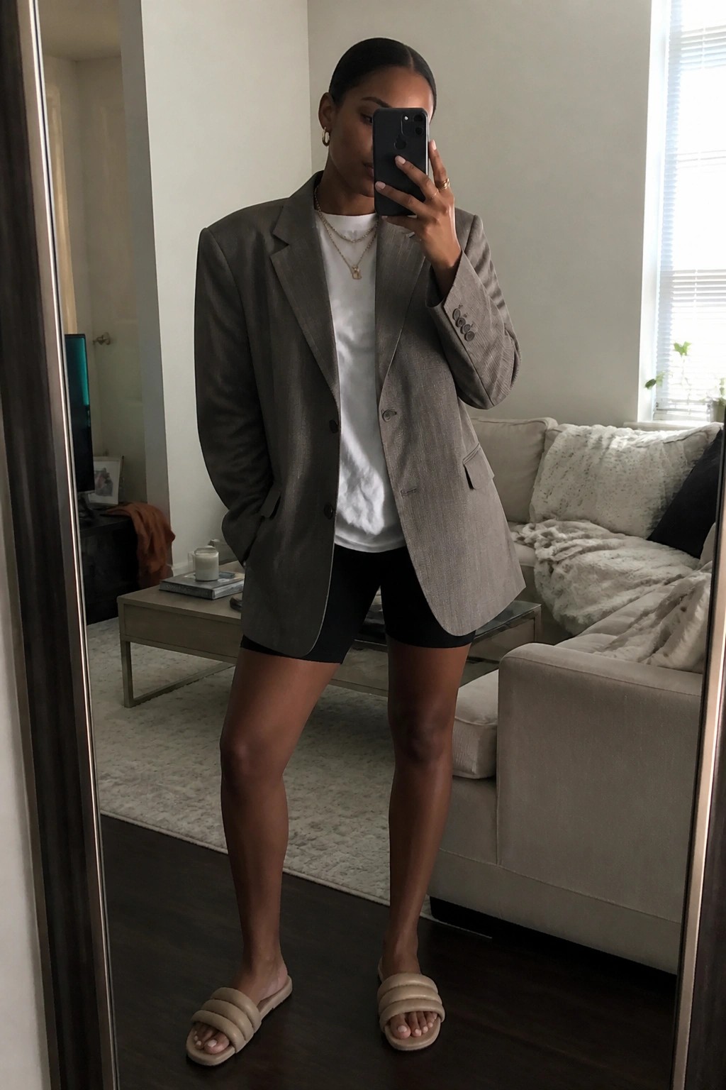 Woman in gray blazer, white tee, and black shorts takes mirror selfie in living room