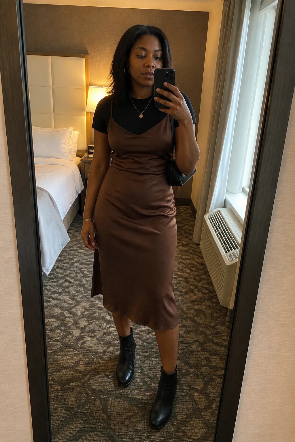 Woman in brown satin dress and black boots taking mirror selfie in hotel room