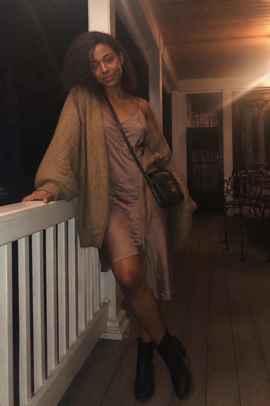 Woman in satin slip dress and chunky cardigan on wooden porch at night.