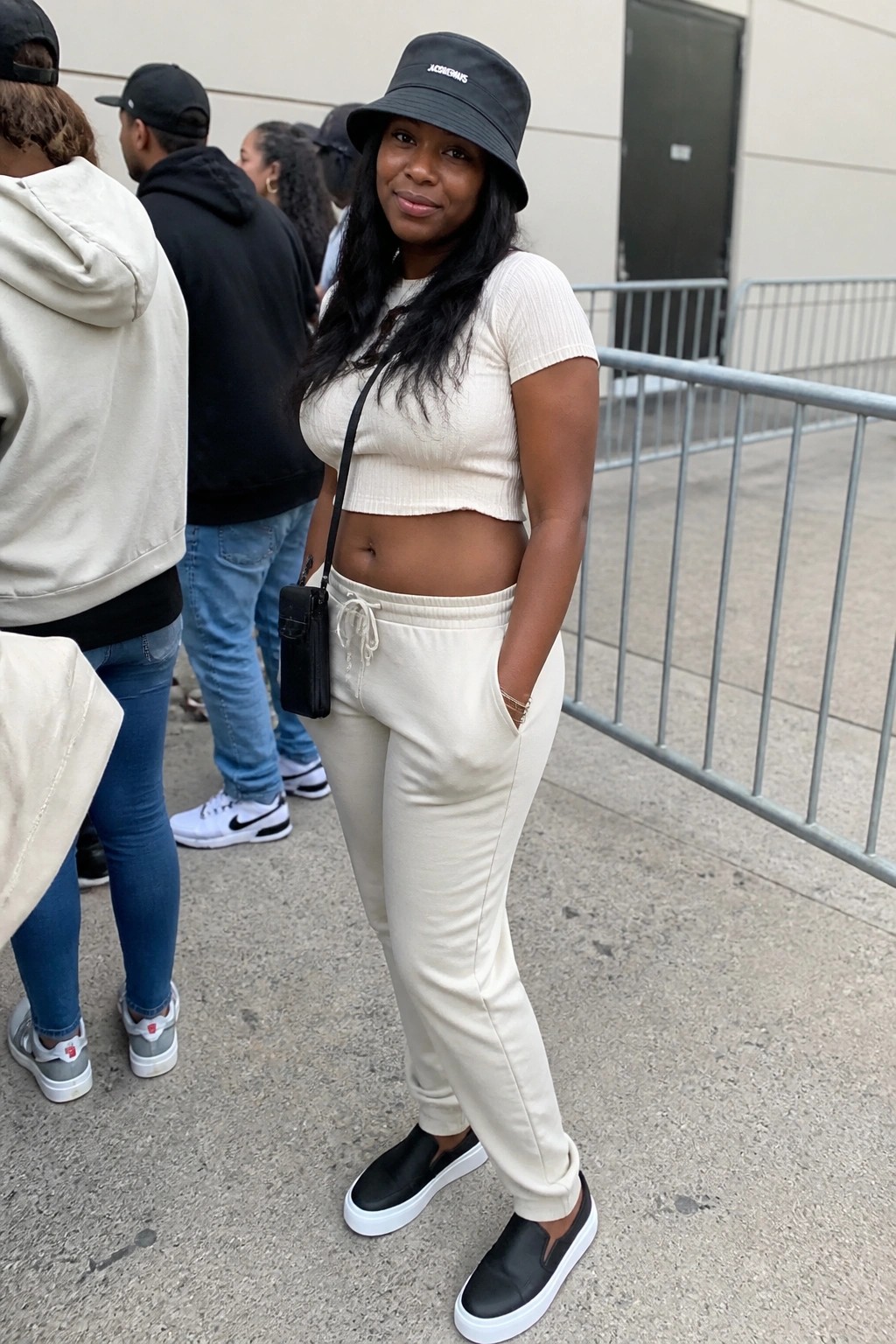 A plus size Black woman wearing a cream cropped top and jogger pants set with a black bucket hat and black sneakers at a concert venue.