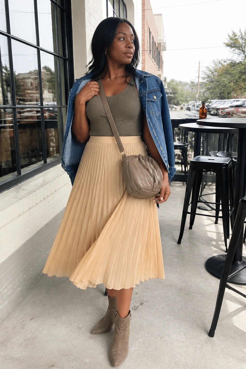 Woman in olive top, beige pleated skirt, denim jacket, and crossbody bag outdoors.