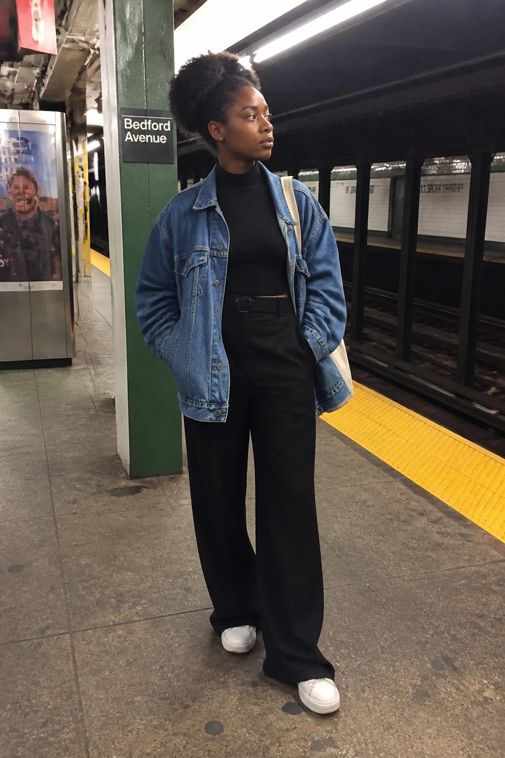Woman in blue denim jacket and black wide-leg pants on subway platform.