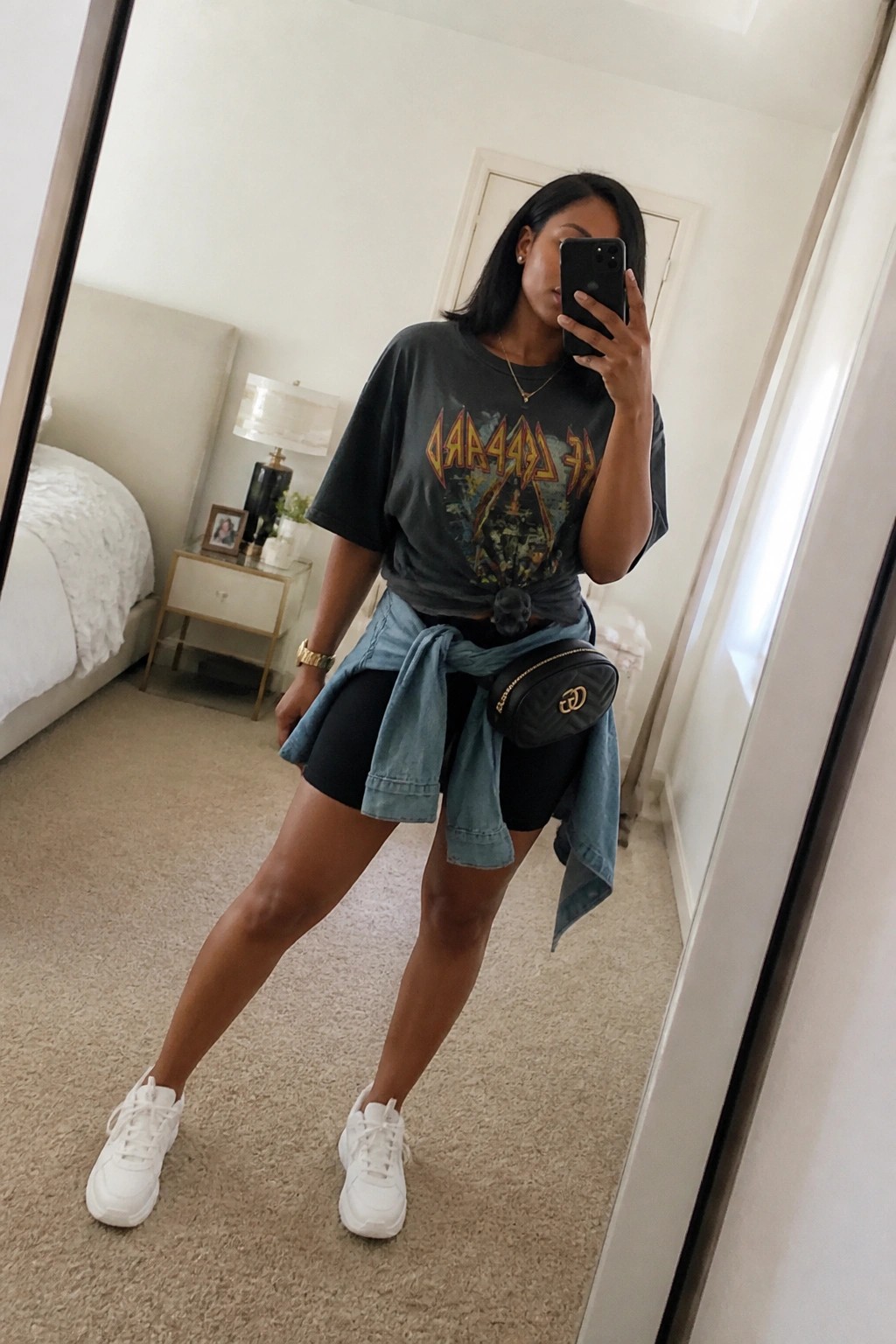 Woman in graphic tee and black shorts with denim tied at waist taking mirror selfie
