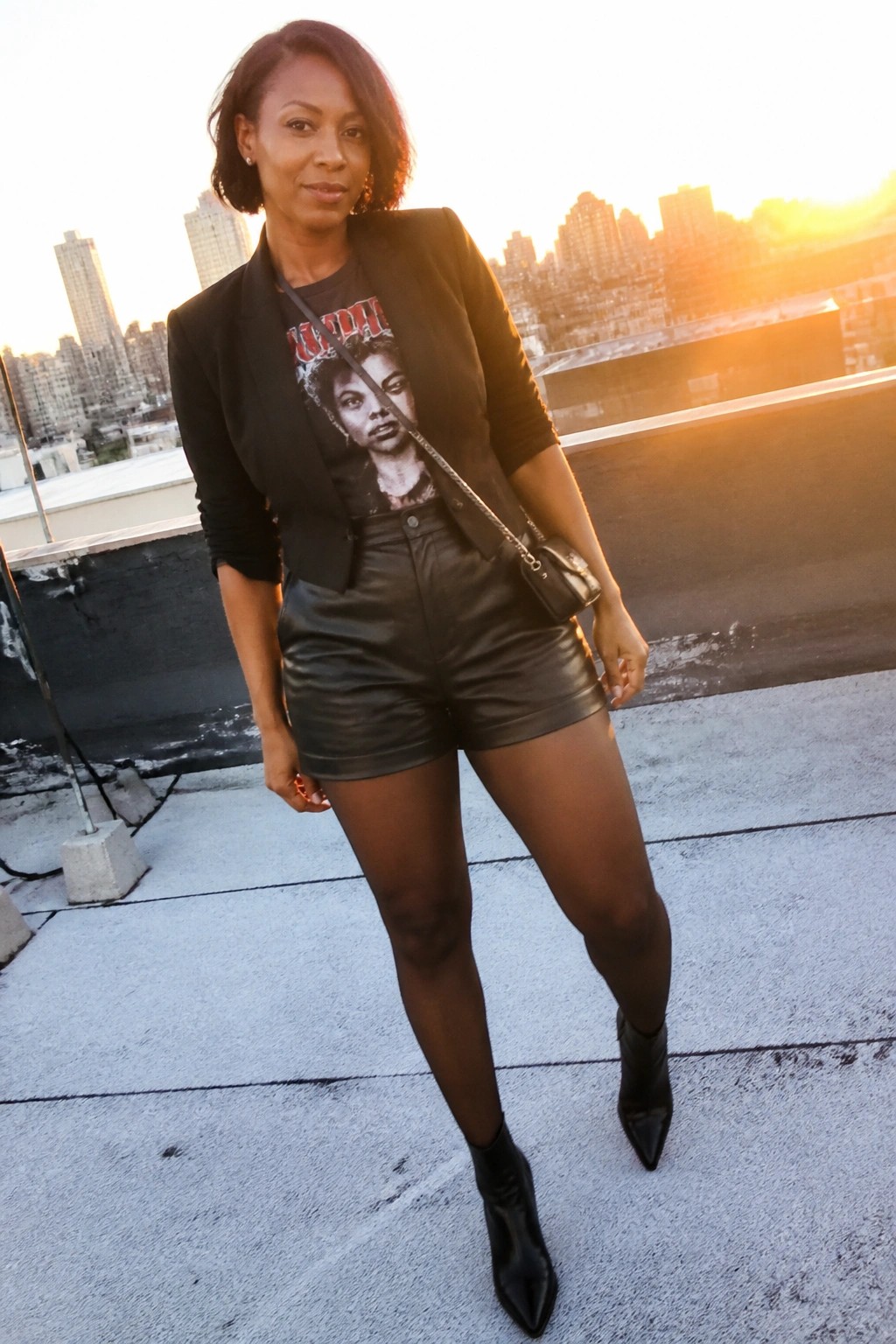 Woman in black blazer, leather shorts, and boots on city rooftop at sunset