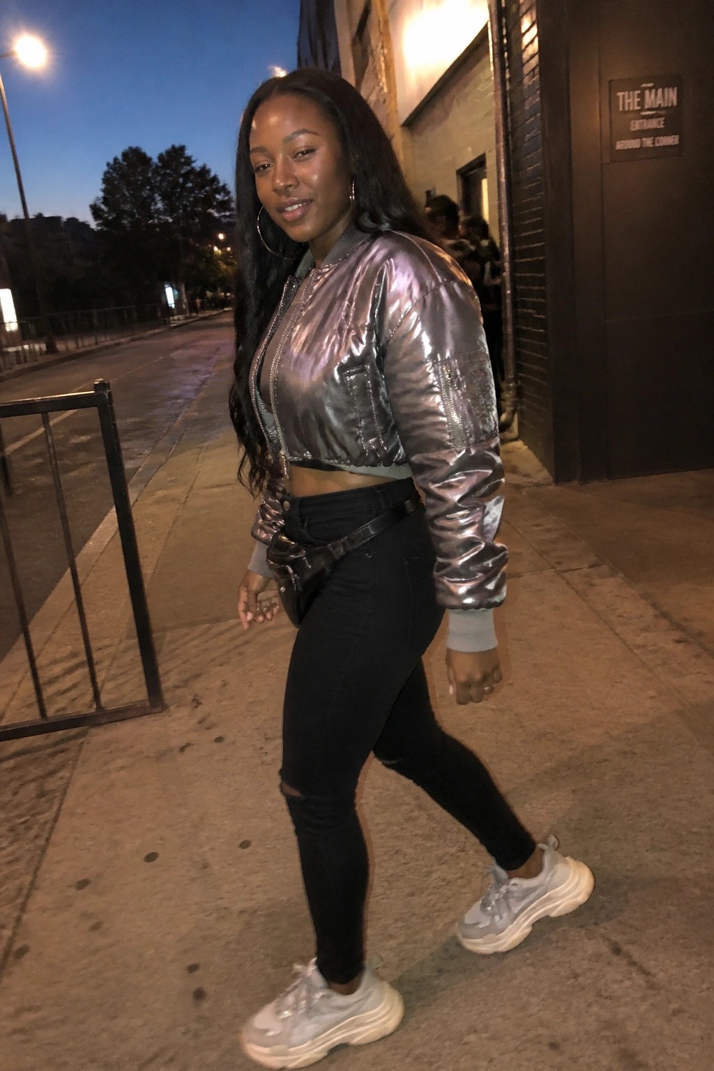 Woman in metallic silver bomber jacket and black jeans on nighttime city sidewalk