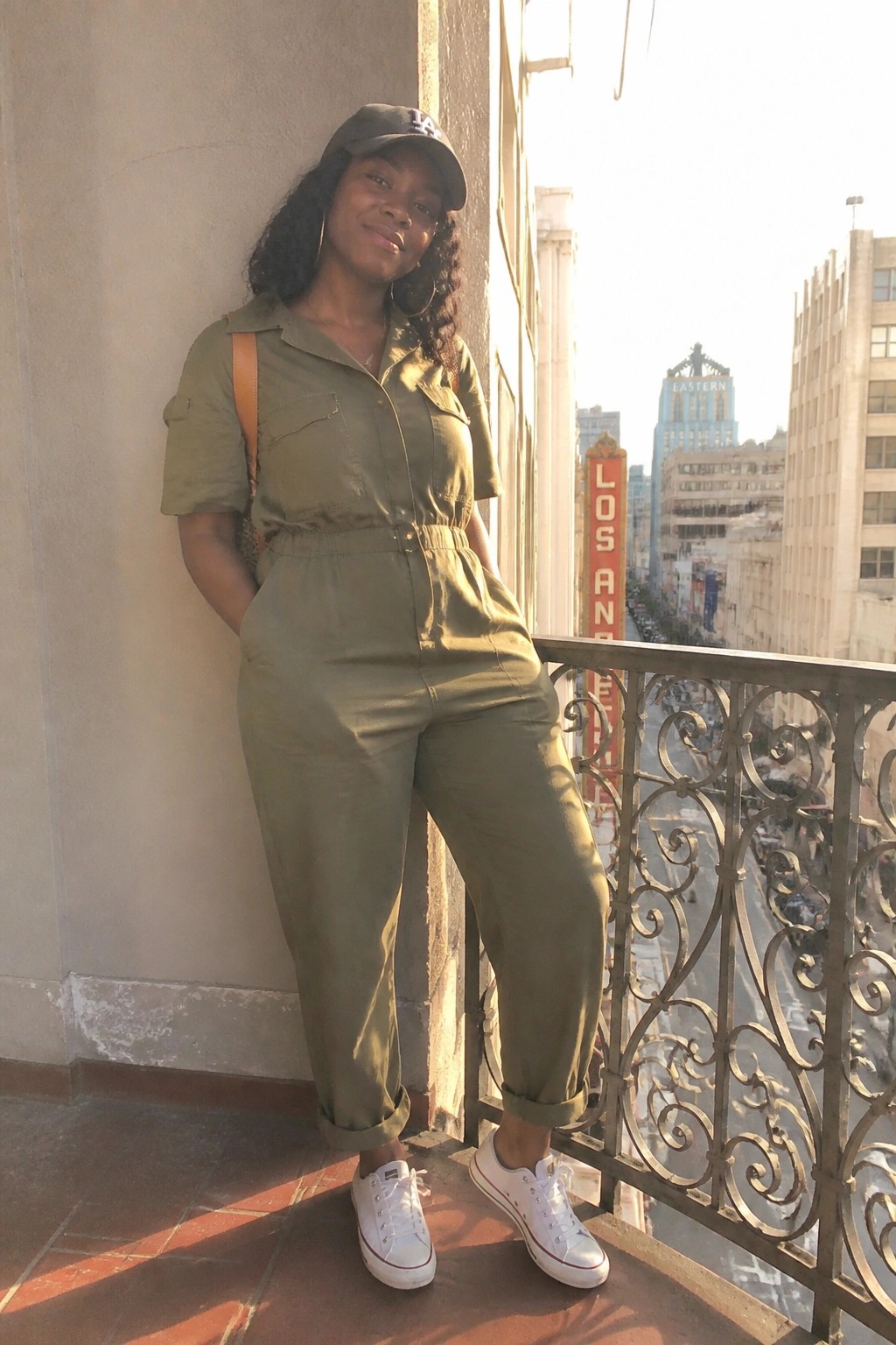 Woman in olive jumpsuit, cap, and white sneakers on sunny city balcony