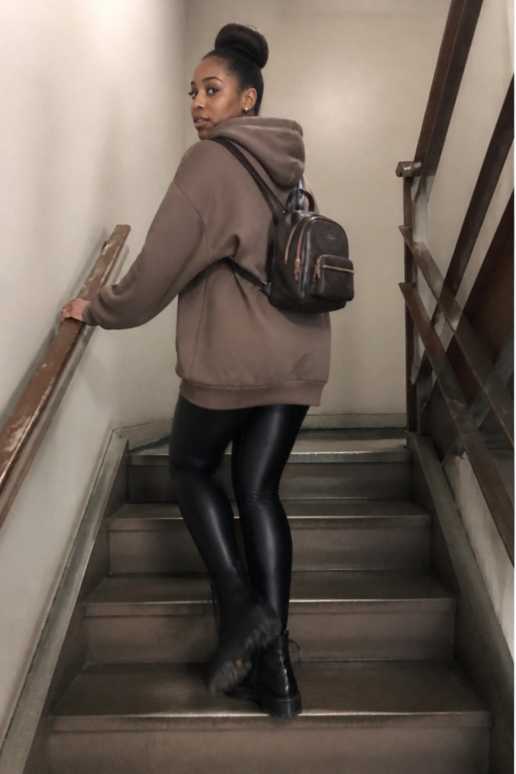 Woman in taupe oversized hoodie, black shiny leggings, and black combat boots standing on indoor stairs with a small brown backpack.