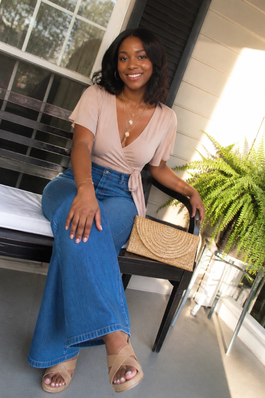 Smiling woman in beige wrap top and blue jeans on porch with woven clutch.