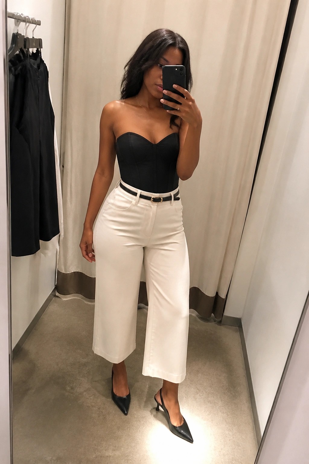 Woman in black strapless corset top, white wide-leg cropped pants with black belt, black pointed heels, taking mirror selfie in fitting room.