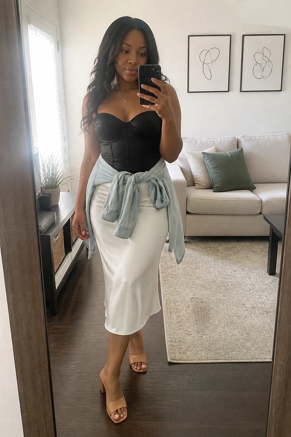Woman in black strapless corset top, white satin midi skirt, light blue denim shirt tied at waist, nude block heel sandals, and thin gold necklace taking a mirror selfie.