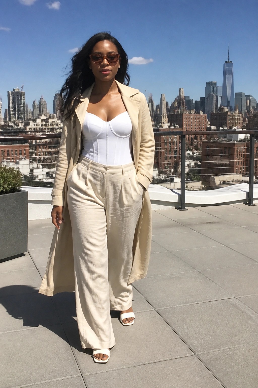 Woman in beige trench coat over white corset on NYC rooftop with skyline view