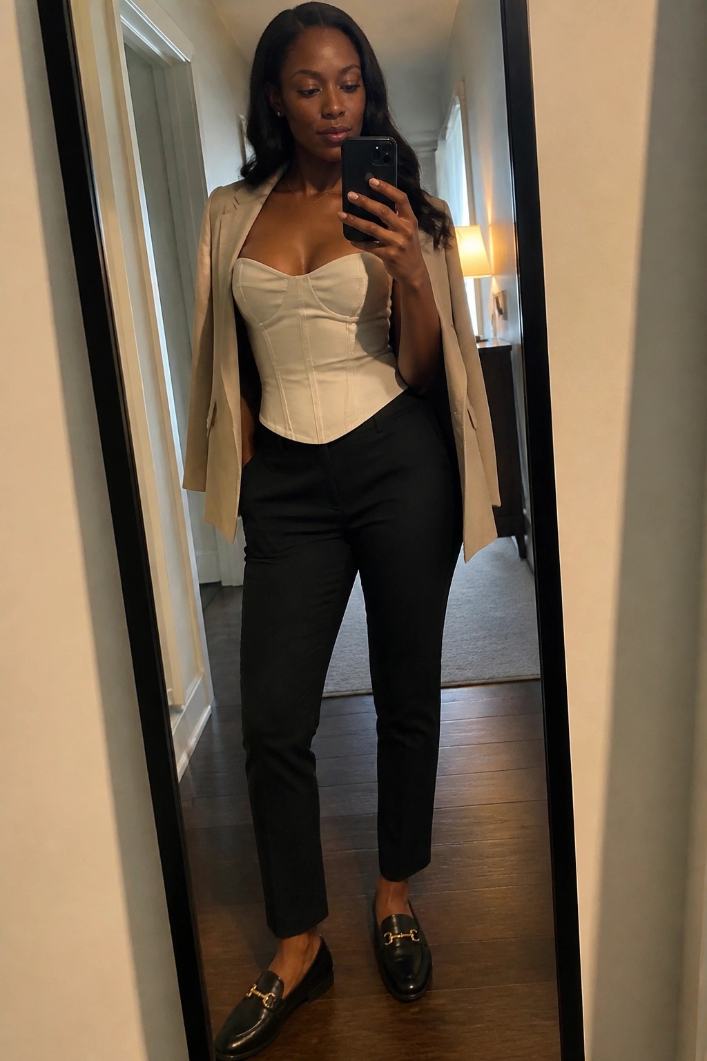 Woman in white corset, beige blazer, and black pants taking mirror selfie