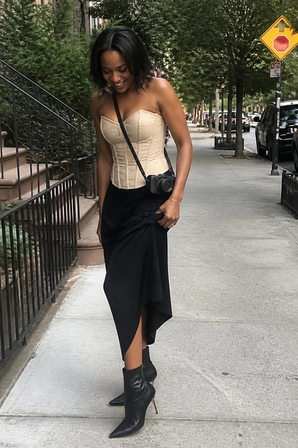 Smiling woman in beige corset top and black skirt carries camera on city sidewalk.