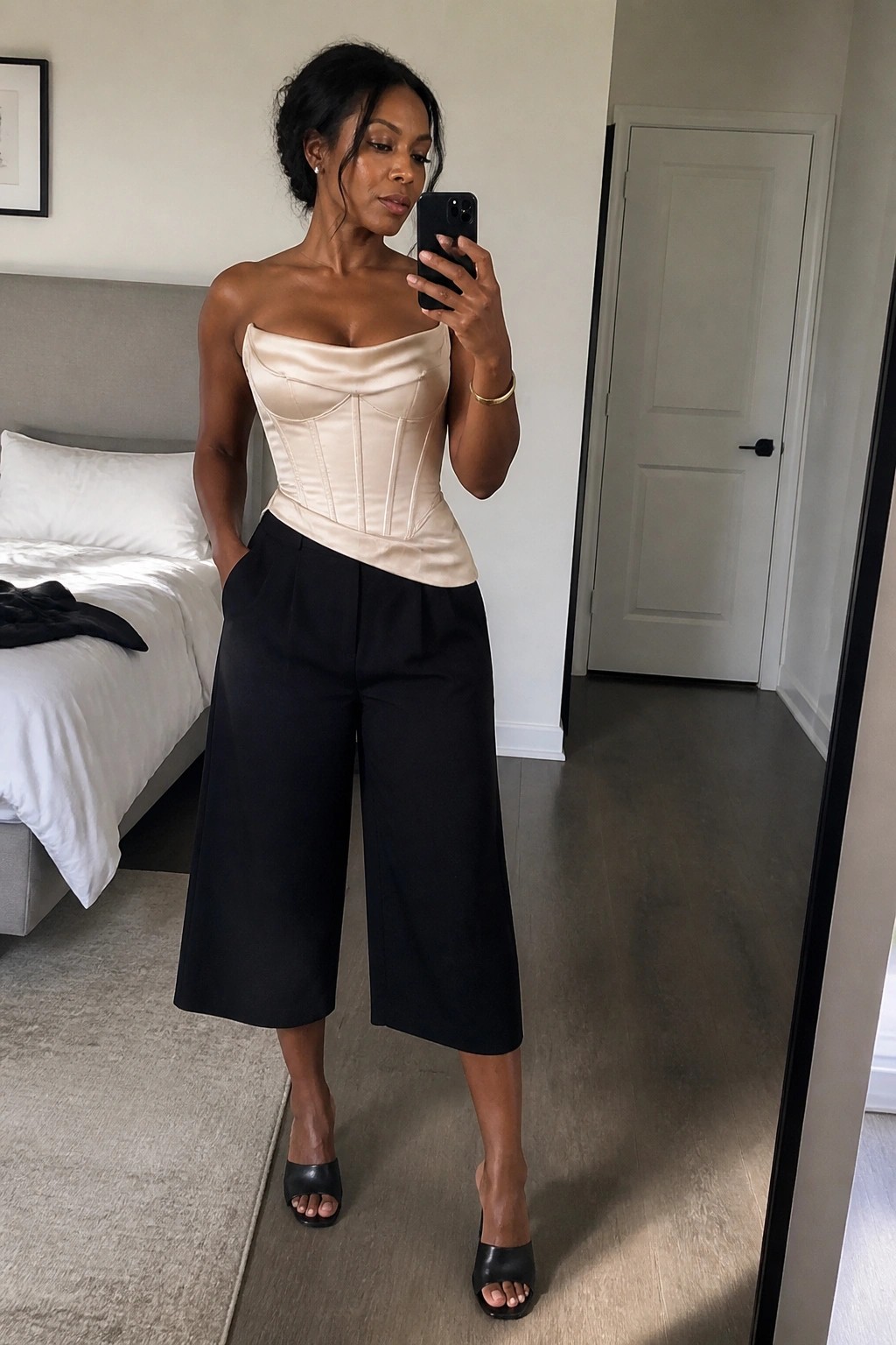 Woman in beige strapless corset top and black wide pants taking mirror selfie in bedroom