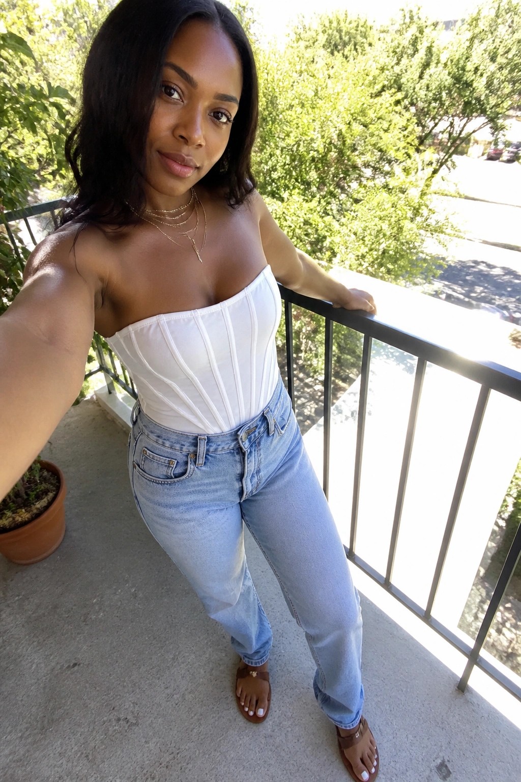 Woman in white strapless corset and blue jeans posing for selfie on sunny balcony