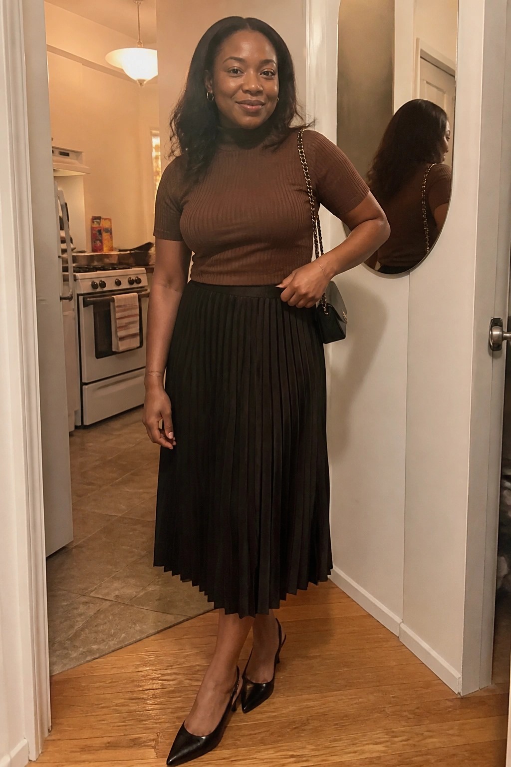 Woman in brown turtleneck and black pleated skirt with chain-strap bag in doorway.