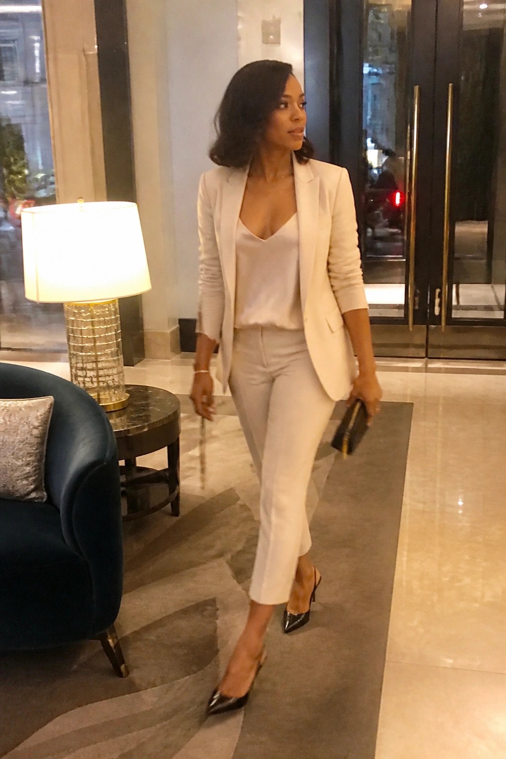 Woman in cream pantsuit, satin camisole and black heels walks in luxury lobby