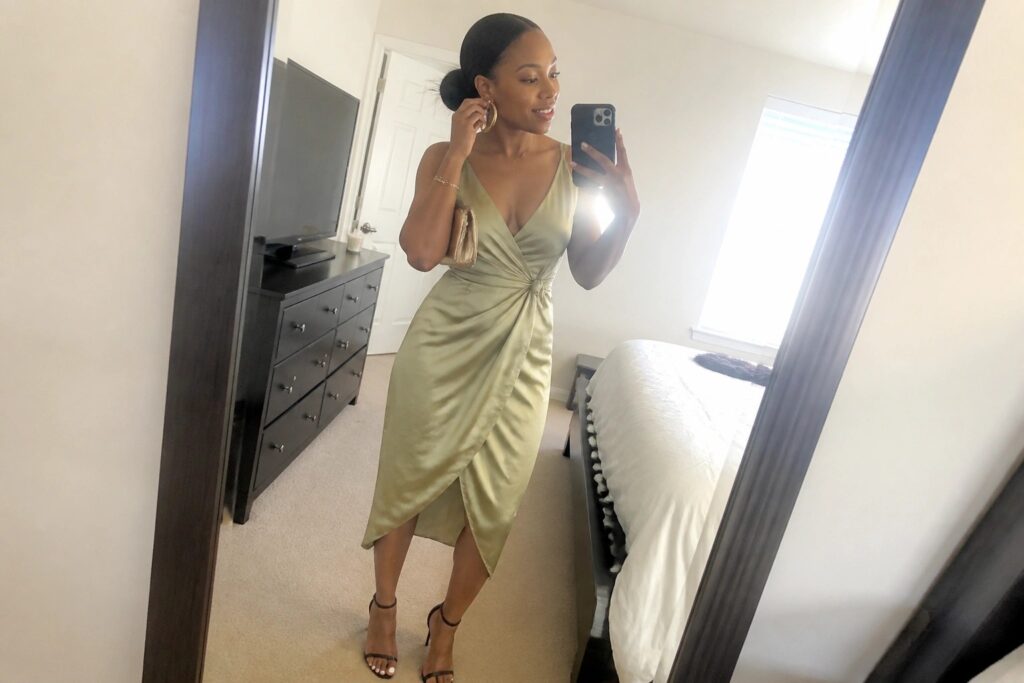 24 Elegant Girls’ Night Dinner Outfit Ideas for Black Women A woman wearing a sage green satin wrap midi dress with puff sleeves and a side knot, paired with black strappy heels and gold hoop earrings, taking a mirror selfie in a bedroom.