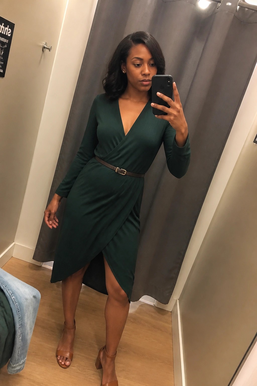 Black woman wearing dark green long sleeve wrap dress with brown belt and matching block heel sandals taking a mirror selfie indoors.