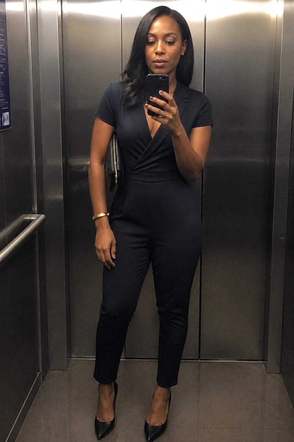 Woman wearing a navy wrap-front jumpsuit with short sleeves, black pointed-toe heels, and holding a black phone and clutch in an elevator.