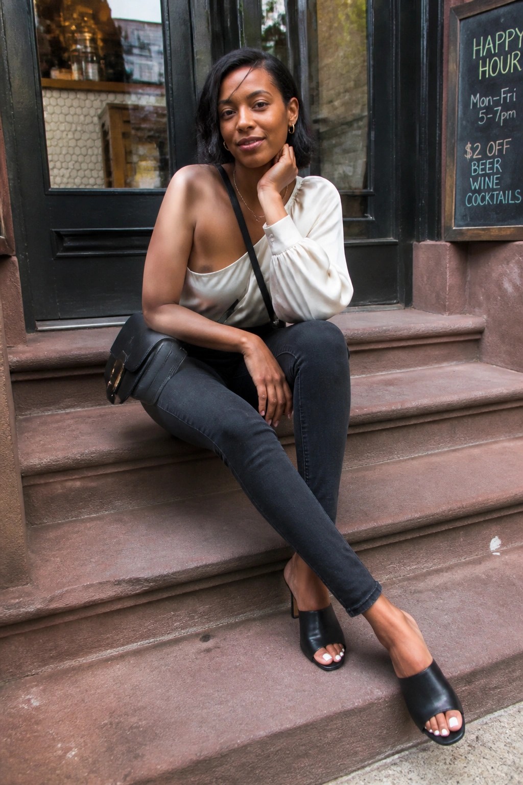 Woman in cream one-shoulder top and black jeans seated on stone steps