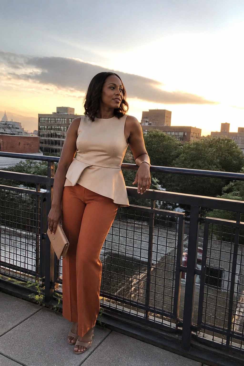 Woman in beige peplum top and orange pants leaning on city balcony at sunset