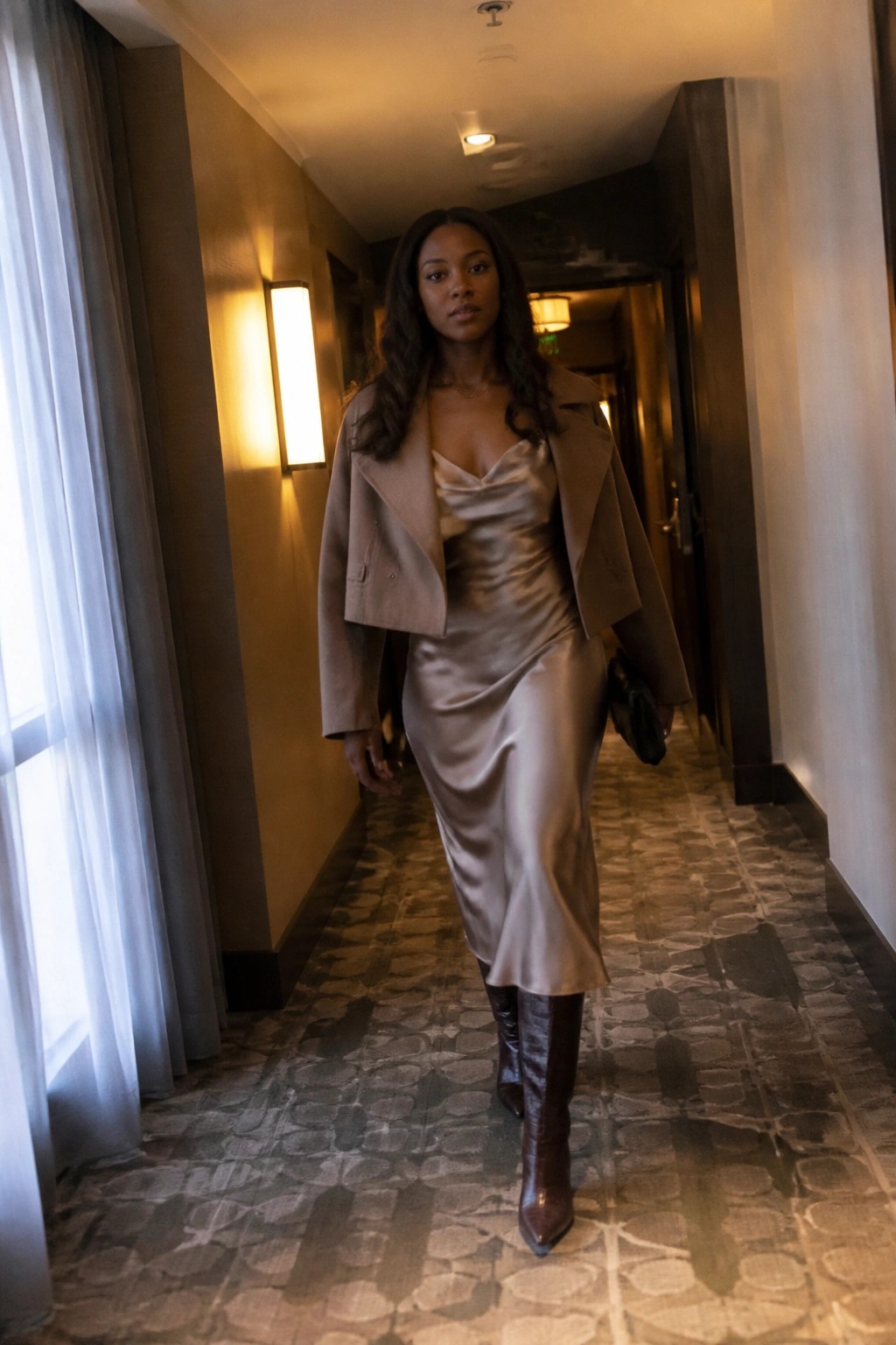 Woman in beige satin dress, cropped jacket, and brown knee-high boots in hotel hallway.