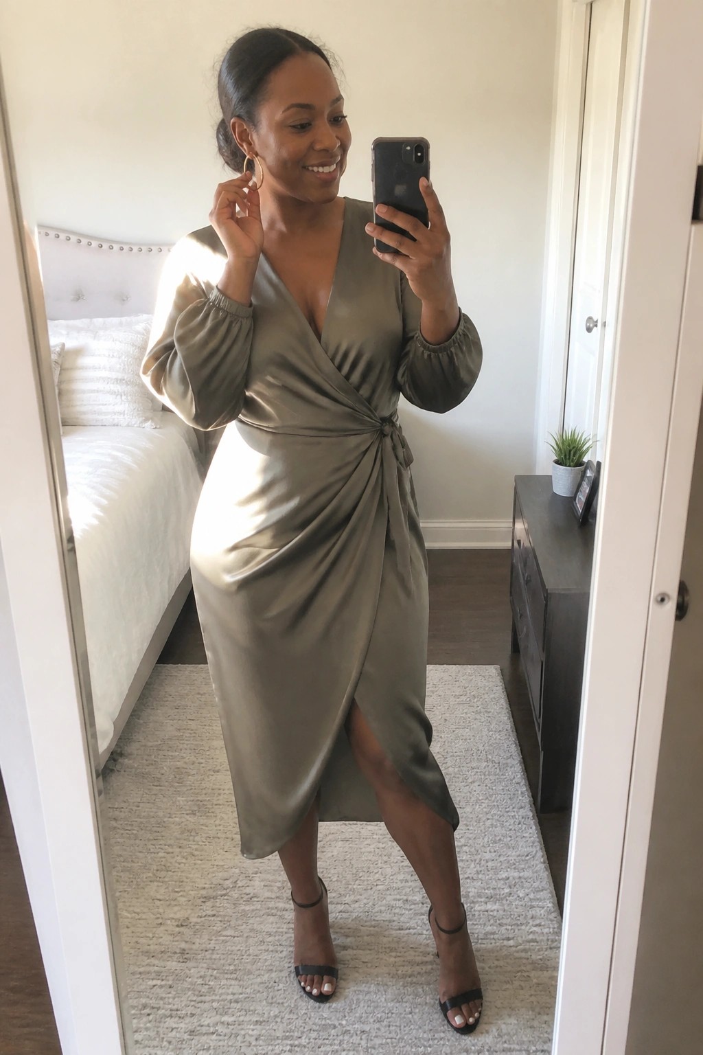 A woman wearing a sage green satin wrap midi dress with puff sleeves and a side knot, paired with black strappy heels and gold hoop earrings, taking a mirror selfie in a bedroom.