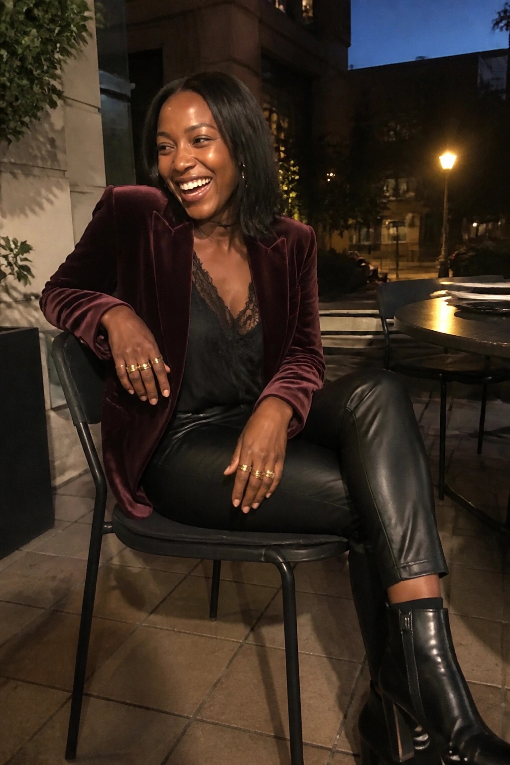 A Black woman seated in a burgundy velvet blazer over a black lace camisole with black leather pants and black heeled boots wearing gold rings.