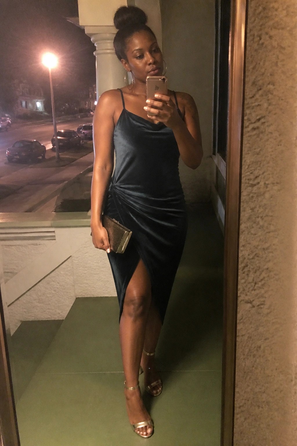 Woman in dark velvet dress taking selfie on porch at night with gold heels