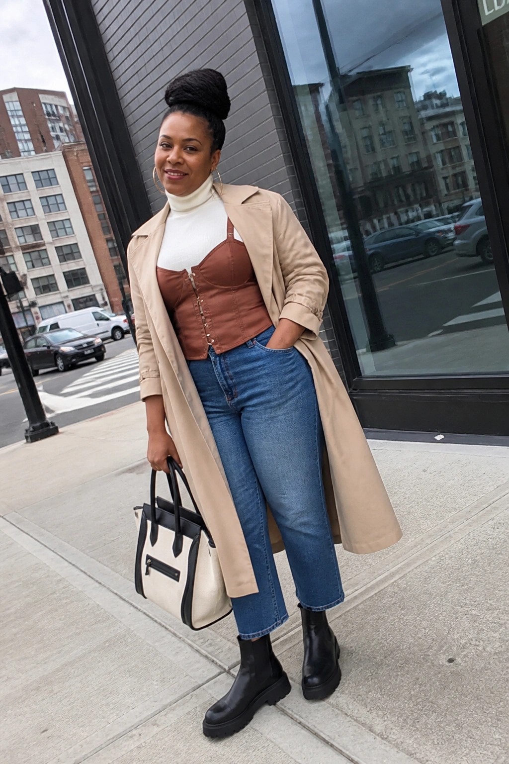 Woman in beige trench coat, brown corset, white turtleneck, blue jeans on sidewalk.
