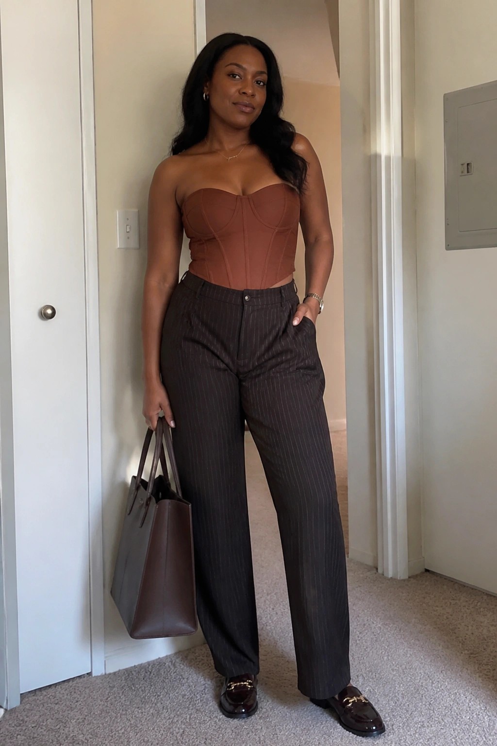 Woman in brown corset top and pinstripe pants holding leather tote bag indoors.