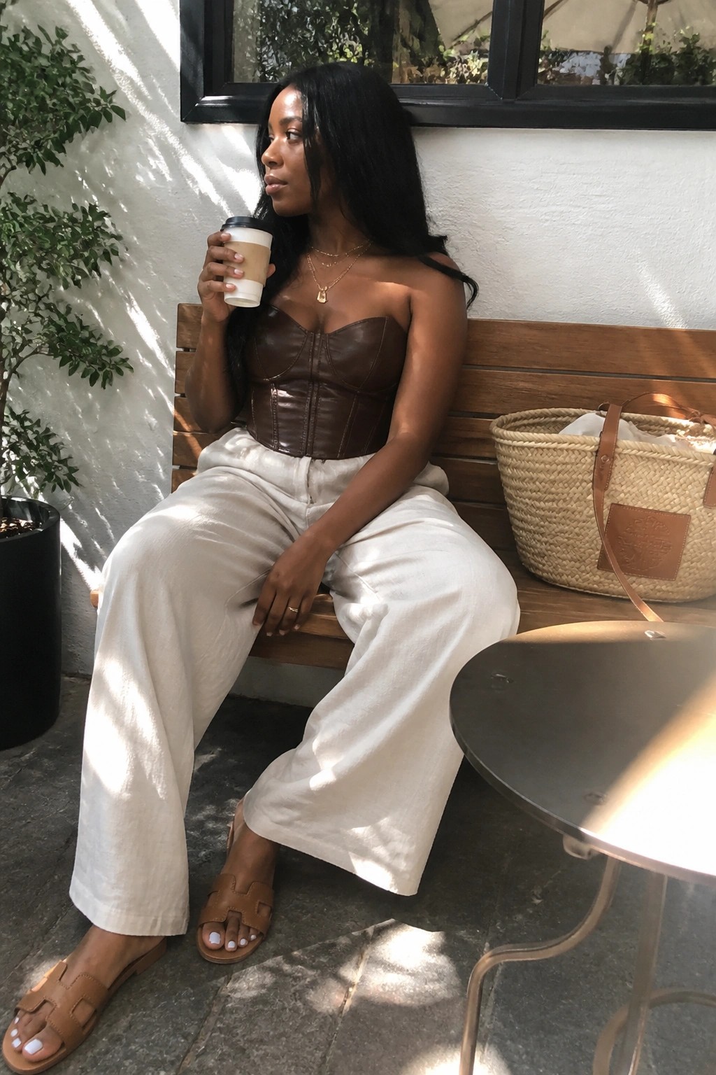 Woman in brown strapless corset top and white wide leg pants with brown slide sandals sitting on a bench holding a coffee cup.