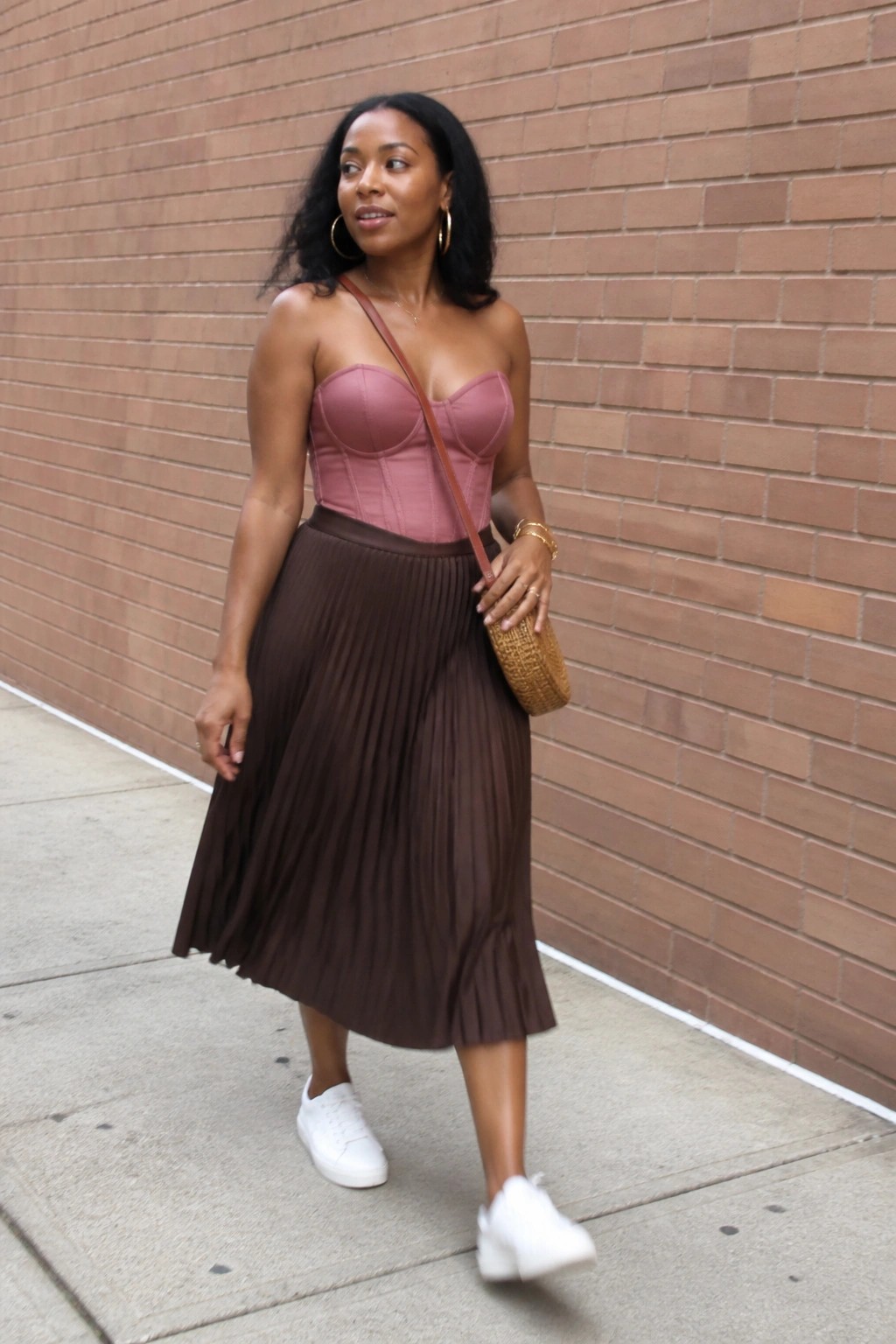 Woman in pink corset top and brown pleated skirt with woven bag by brick wall