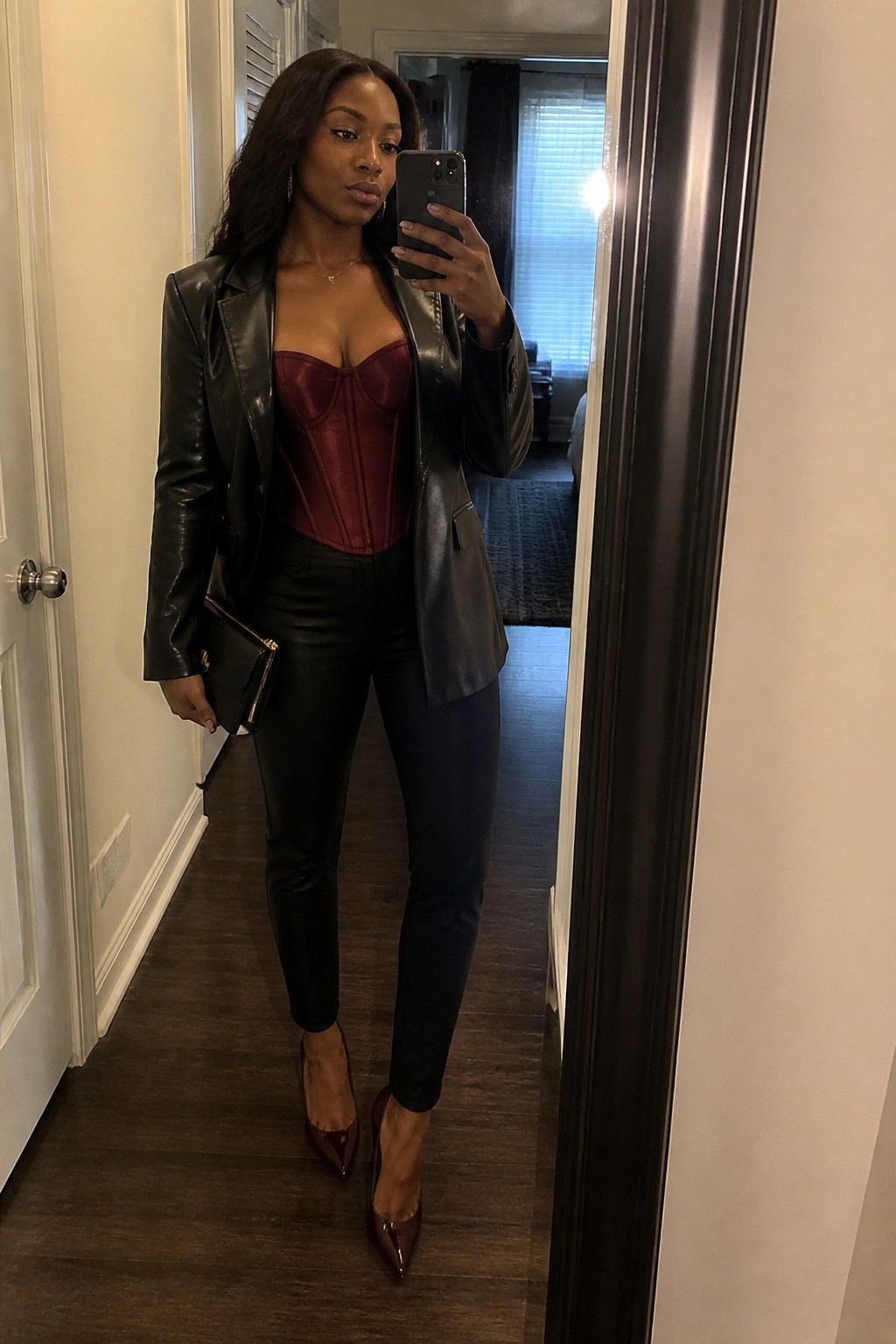 Woman in black leather jacket over maroon corset posing for mirror selfie in hallway