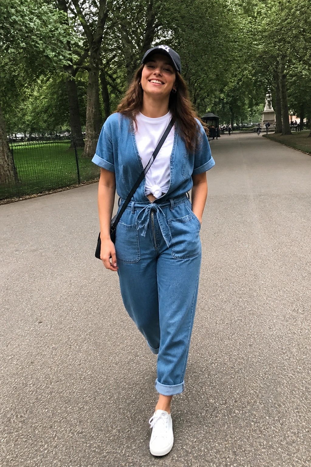 Smiling woman in blue denim jumpsuit, cap, and white sneakers on park path