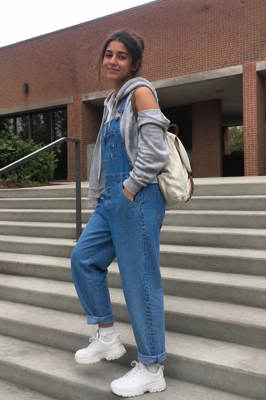 Young woman in blue denim overalls over gray hoodie on concrete steps with backpack