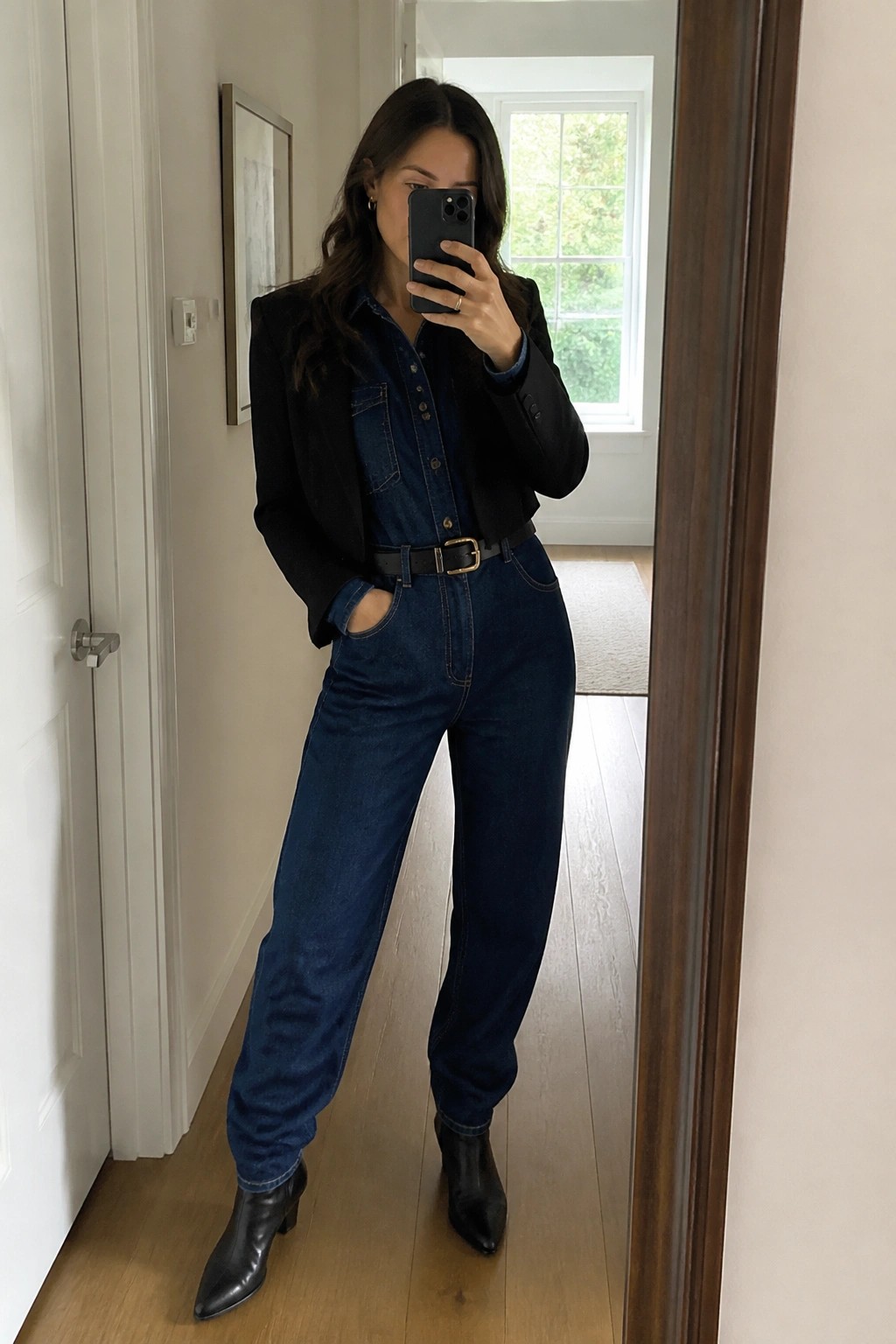 Woman in blue denim jumpsuit and black blazer taking mirror selfie with phone.