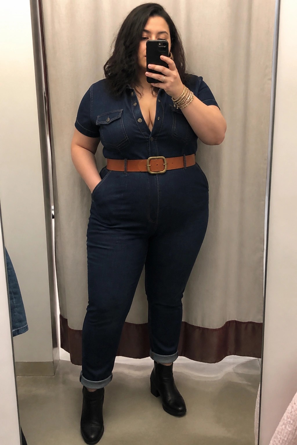 Woman in denim jumpsuit with brown belt taking mirror selfie in fitting room