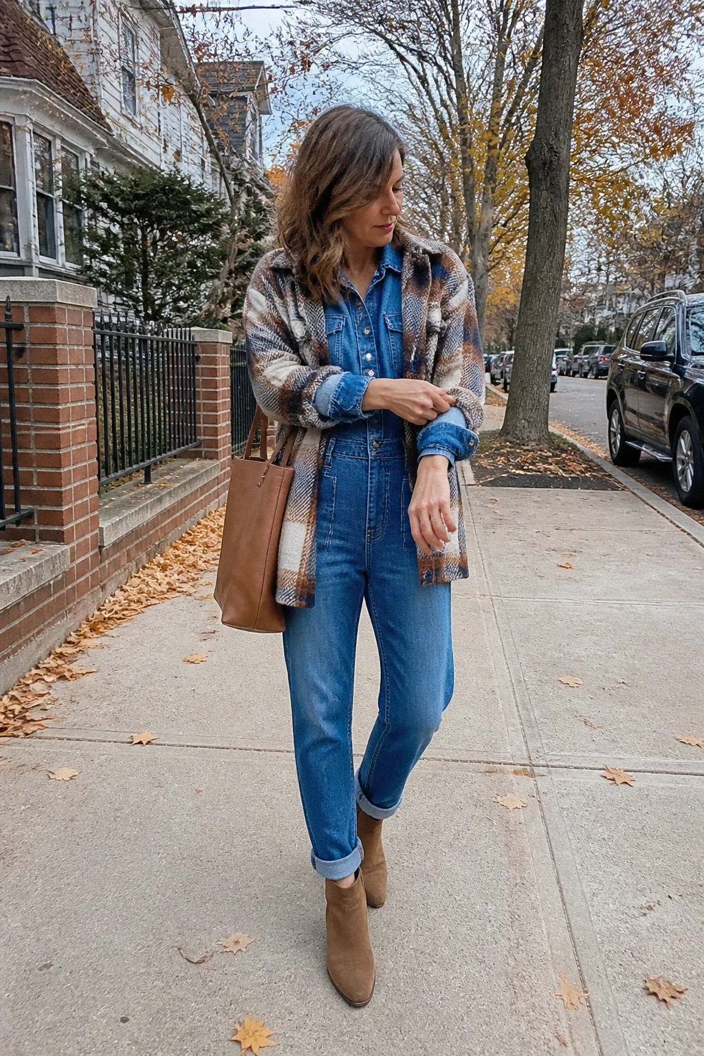 Woman in blue denim jumpsuit, plaid coat, and brown boots on autumn sidewalk.