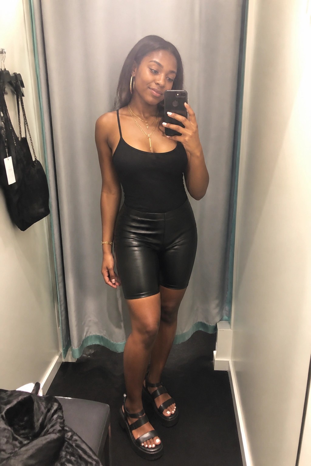 Woman in black bodysuit and shiny shorts taking mirror selfie in fitting room