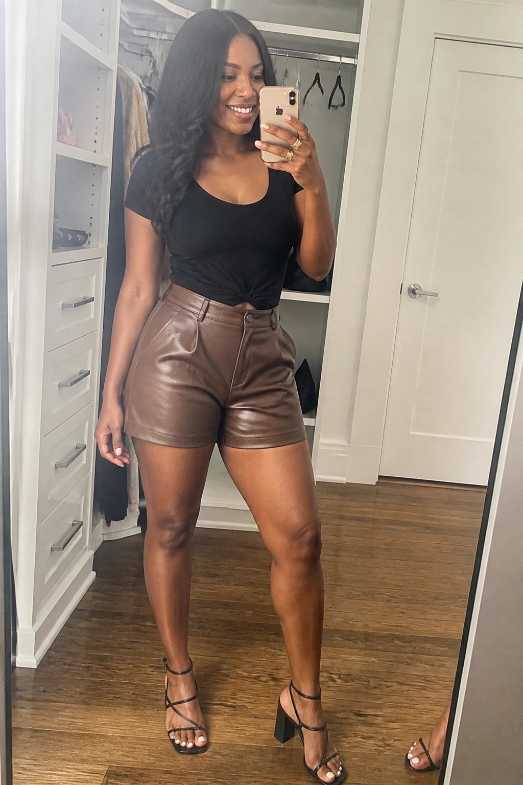 Woman wearing a black knotted short sleeve top with brown leather shorts and black strappy block heel sandals while taking a mirror selfie in a closet area