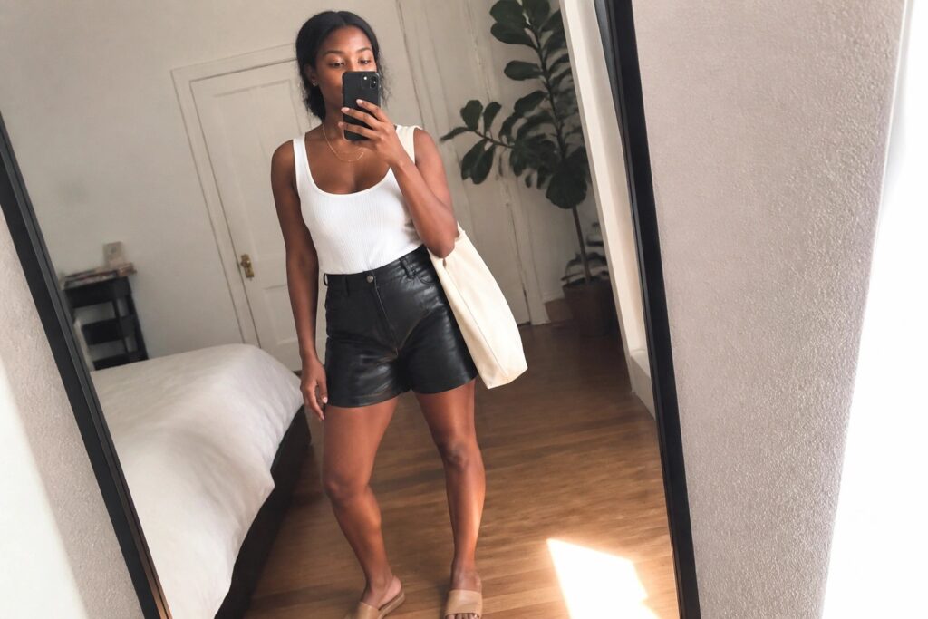 Woman in white tank top and black leather shorts holding phone and tote bag in bedroom